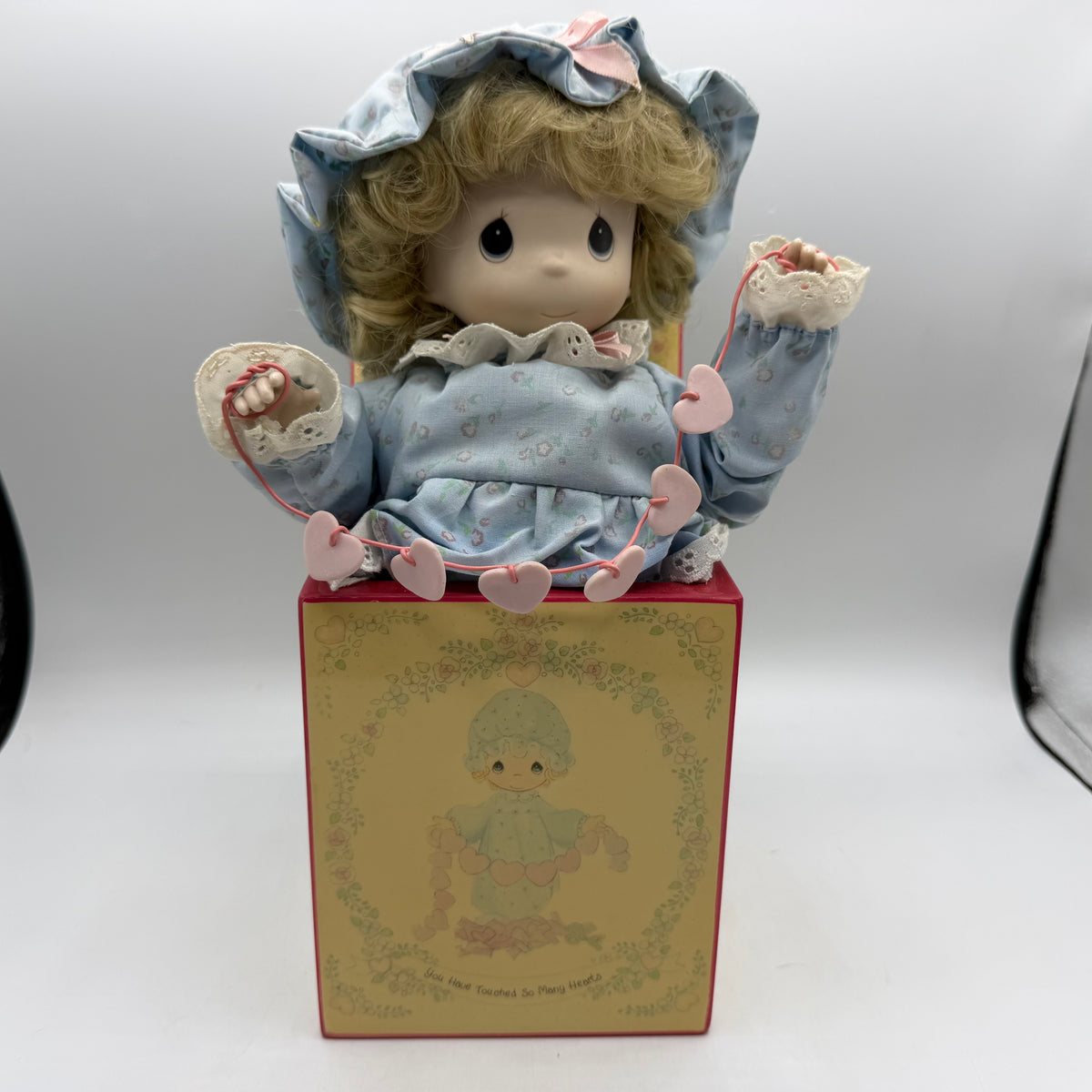 A vintage Precious Moments doll with blonde curly hair, wearing a light blue dress and matching bonnet with pink accents, posed on top of its illustrated display box against a white studio background.