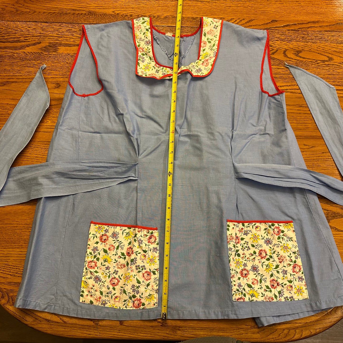 Gray apron with floral pockets and trim on a wooden surface, measured by a tape measure.