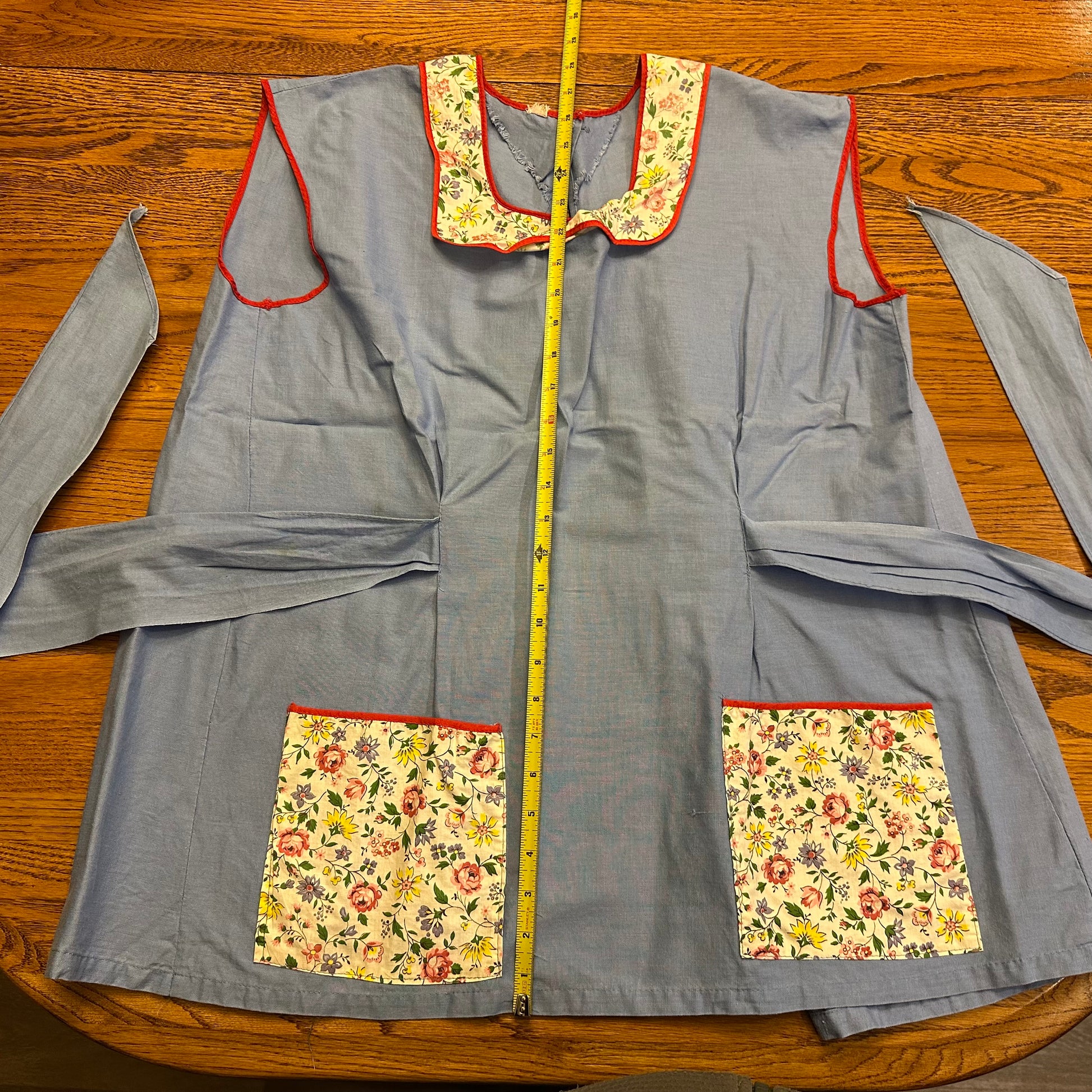 Gray apron with floral pockets and trim on a wooden surface, measured by a tape measure.