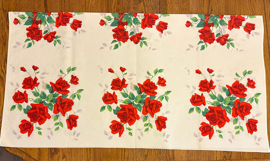 A rectangular cream-colored tablecloth featuring a repeating pattern of red roses and green leaves, laid flat on a wooden floor.